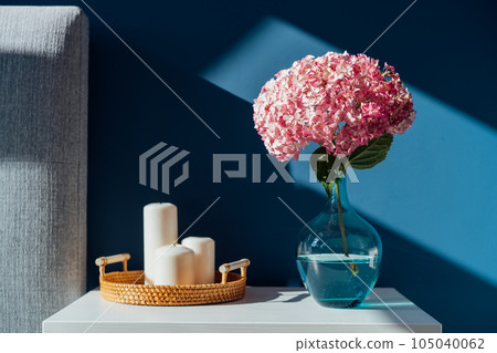 Stylish modern bedroom in dark colors. Cozy interior with navy blue walls, home decor. Bed with grey fabric headboard, white bedside table, vase with pink hydrangea flower, candles on wicker tray. 105040062