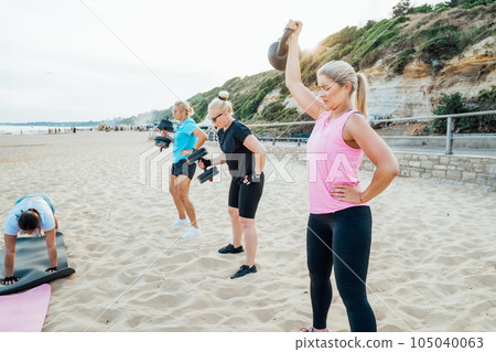 Active women of various ages doing fitness workouts in class exercise with coach on the beach. Ladies working with dumbbells and doing plank. Sport for health and wellbeing. Active lifestyle 105040063