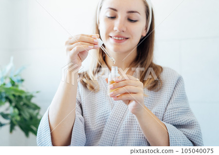 Close up young woman in bathrobe holding mockup bottle and dropper with hyaluronic acid serum in her hands. Skin hydrating. Cosmetic spa procedures. Beauty self-care at home. Selective focus 105040075