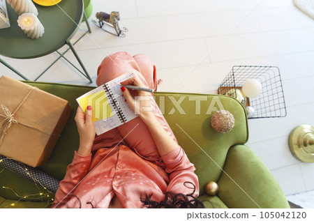 Top view of woman making notes in notebook while sitting on sofa at home during winter holidays Top view of woman making notes in notebook while sitting on sofa at home during winter holidays 105042120