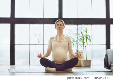 Meditation during pregnancy. Young calm tranquil pregnant woman doing yoga at home, meditating and practicing mindfulness, expectant mother sitting in lotus pose with closed eyes Meditation during pregnancy. Young calm tranquil pregnant woman doing yoga at home, meditating and practicing mindfulness, expectant mother sitting in lotus pose with closed eyes 105042149