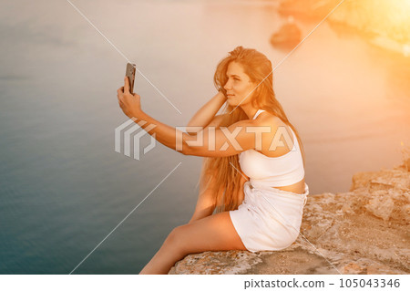 Selfie woman sea. white tank top, and shorts captures a selfie shot with her mobile phone against the backdrop of a serene beach and blue sea. 105043346