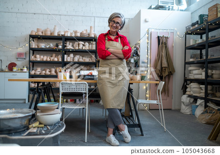 Happy senior woman successful self-employed potter wearing apron behind table with pottery tools. 105043668