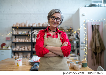 Happy senior woman successful self-employed potter wearing apron behind table with pottery tools. 105043672