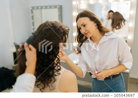 Medium shot of focused makeup artist applying eyeshadow powder to eyelids of unrecognizable woman in beauty salon. Hairstylist doing stylish hairdo. Concept of four-handed makeup and styling. 105043697
