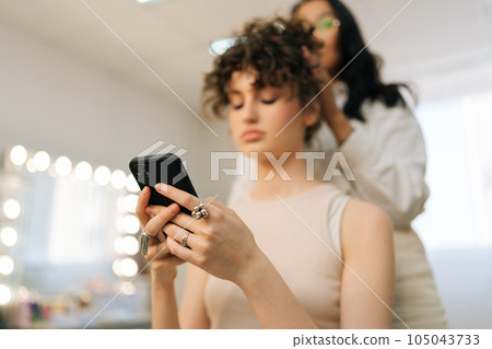 Closeup low-angle view of young woman client sitting in chair in beauty salon using phone while master hairdresser giving beauty salon service and treatment. Hairstylist doing stylish hairdo. 105043733