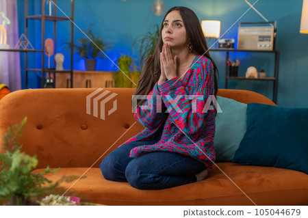 Young woman girl praying sincerely with folded arms asking God for help begging apology on home sofa Young woman girl praying sincerely with folded arms asking God for help begging apology on home sofa 105044679