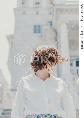 Wind hair style. A portrait of a woman outdoors, her shoulder-length brown hair blowing in the wind. Dressed in a white shirt against a light building. 105044809