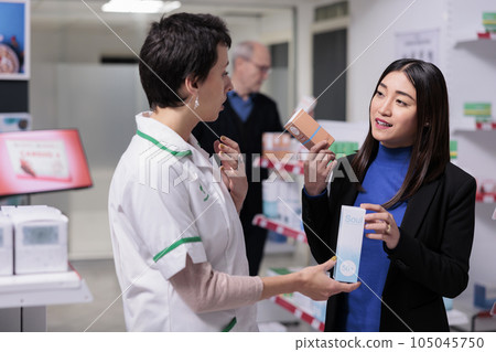 Pharmacist recommending young asian woman sunblock cream in drugstore. Customer holding skin care products packages, choosing between two sunscreen lotions and consulting with pharmacy assistant 105045750