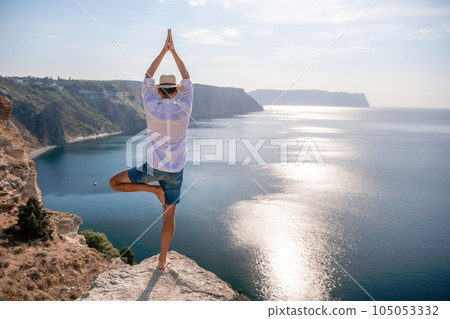 A digital nomad, a man in a hat, a businessman does yoga on the rocks by the sea at sunset time, does a business operation online from a distance. Remote work on vacation. 105053332