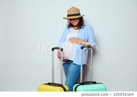 Pregnant woman in straw hat with colorful suitcases, strokes her belly, smiles enjoying her travel abroad in pregnancy 105056164
