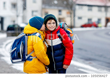 Two little kids boys of elementary class walking to school during snowfall. Happy children having fun and playing with first snow. Siblings and best friends with backpack in colorful winter clothes. 105056427