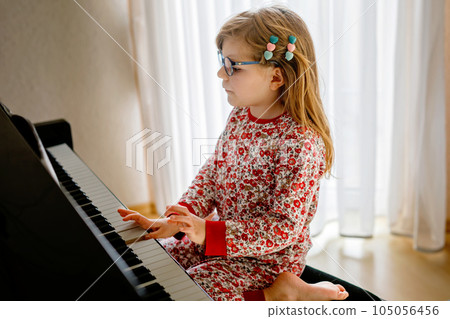 Little happy girl playing piano in living room. Cute preschool child with eye glasses having fun with learning to play music instrument. Little happy girl playing piano in living room. Cute preschool child with eye glasses having fun with learning to play music instrument. 105056456