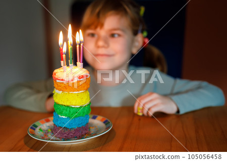 Happy little preschool girl celebrating birthday. Closeup of child with homemade rainbow cake, indoor. Happy healthy toddler blowing six candles on cake. Selective focus on cake 105056458