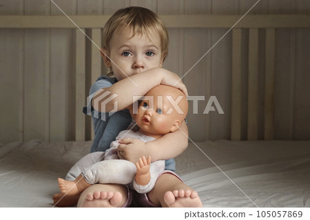 Girl and Baby Doll. Little Girl Playing on a White Blanket in Bedroom. Front View. Child Holding the Doll Toy. Family Relationship Concept. Kid Toddler is Alone, She Hugging and Cradling Her Toy 105057869