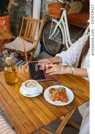 In a summer cafe, a woman surfs the net on a laptop while eating a croissant 105059427