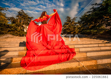 Sunrise red dress. A woman in a long red dress against the backdrop of sunrise, bright golden light of the sun's rays. The concept of femininity, harmony. 105061661