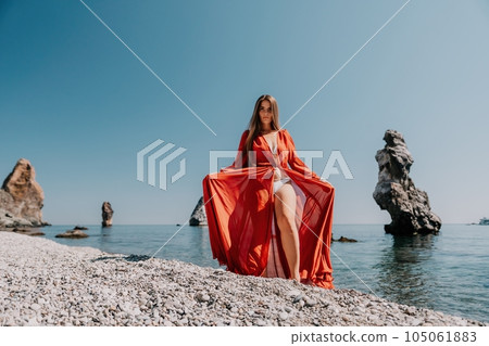 Woman travel sea. Happy tourist in red dress enjoy taking picture outdoors for memories. Woman traveler posing on the rock at sea bay surrounded by volcanic mountains, sharing travel adventure journey Woman travel sea. Happy tourist in red dress enjoy taking picture outdoors for memories. Woman traveler posing on the rock at sea bay surrounded by volcanic mountains, sharing travel adventure journey 105061883