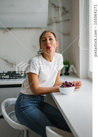 Attractive young woman eats fresh cherries, healthy berries sitting at home in the kitchen and fooling around Attractive young woman eats fresh cherries, healthy berries sitting at home in the kitchen and fooling around 105064271