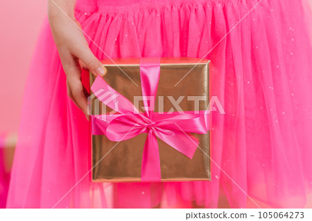 Close up of a girl holding a gift box with a pink silk ribbon, giving a gift for a holiday. Merry Christmas, Happy New Year, Valentine's Day, holiday, festive, Birthday, surprise concept. 105064273