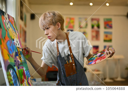 Concentrated young gay male dressed in a blue dirty apron holding a palette and painting on canvas in art studio Concentrated young gay male dressed in a blue dirty apron holding a palette and painting on canvas in art studio 105064888