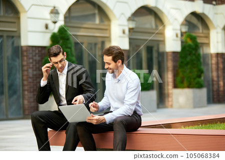 Two businessmen, coworkers sitting on bench outside the office, looking on laptop and working. Analyitcs Two businessmen, coworkers sitting on bench outside the office, looking on laptop and working. Analyitcs 105068384