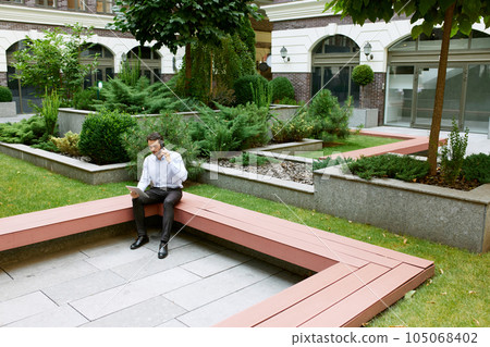Top view. Serious businessman working online with tablet, sitting on bench outside of his office, talking on mobile phone Top view. Serious businessman working online with tablet, sitting on bench outside of his office, talking on mobile phone 105068402