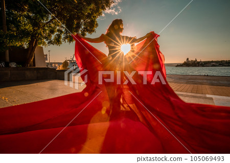 Sunrise red dress. A woman in a long red dress against the backdrop of sunrise, bright golden light of the sun's rays. The concept of femininity, harmony. 105069493