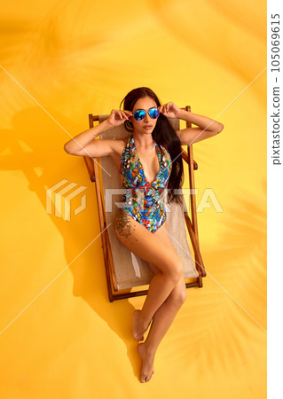 Beautiful young brunette woman in colored swimsuit lies on the lounger on yellow background 105069615