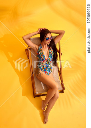 Beautiful young brunette woman in colored swimsuit lies on the lounger on yellow background Beautiful young brunette woman in colored swimsuit lies on the lounger on yellow background 105069616