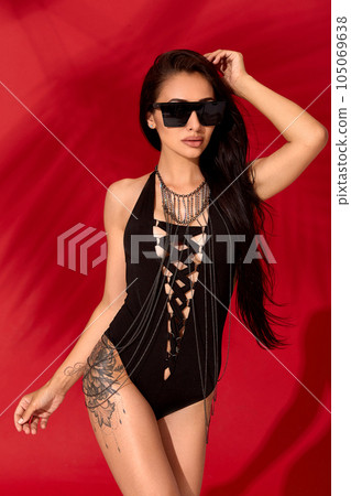 Attractive young and beautiful brunette girl in a black sexy swimsuit posing on red background 105069638