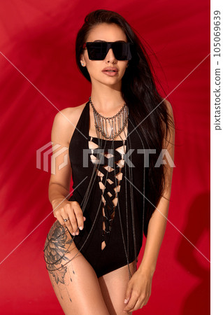 Attractive young and beautiful brunette girl in a black sexy swimsuit posing on red background Attractive young and beautiful brunette girl in a black sexy swimsuit posing on red background 105069639