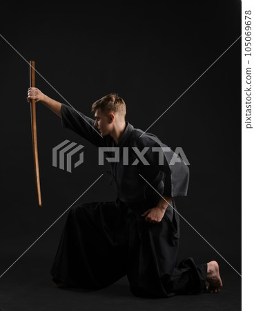 Kendo guru wearing in a traditional japanese kimono is practicing martial art with the shinai bamboo sword against a black studio background. Kendo guru wearing in a traditional japanese kimono is practicing martial art with the shinai bamboo sword against a black studio background. 105069678