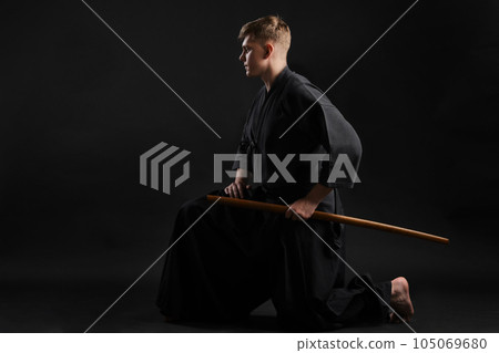 Kendo guru wearing in a traditional japanese kimono is practicing martial art with the shinai bamboo sword against a black studio background. Kendo guru wearing in a traditional japanese kimono is practicing martial art with the shinai bamboo sword against a black studio background. 105069680