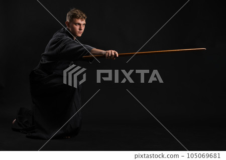 Kendo guru wearing in a traditional japanese kimono is practicing martial art with the shinai bamboo sword against a black studio background. 105069681