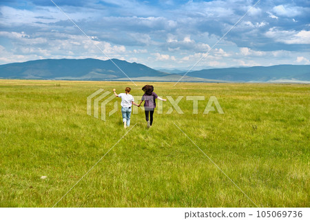 Two girls run through the green grass of the Barguzin Valley in the Republic of Buryatia. View from the back. 105069736