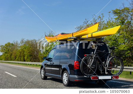 Back view modern black family wagon van car with mounted roof kayak and bike tail carrier driving european highway road against blue sky summer day. Lifestyle travel adventure trip journey concept 105070063