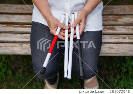 An elderly blind woman sits on a bench in the park. Close-up of female hands with folded tactile cane. 105072948