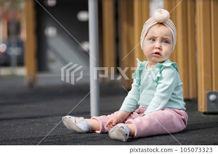 Concerned toddler girl sitting on sunlit playground surface 105073323