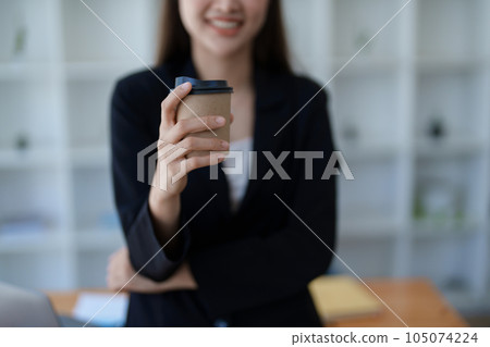 Young minded successful employee business woman in black suit work hold paper cup coffee to go stand at desk with laptop computer Young minded successful employee business woman in black suit work hold paper cup coffee to go stand at desk with laptop computer 105074224
