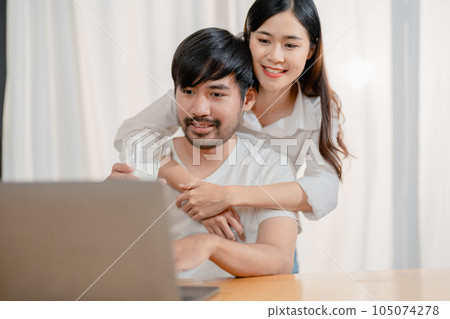 Happy young loving family couple looking at laptop computer screen, reading ecipes. Wife showing embrace to husband to pretty joyful husband, sitting at table in kitchen at home in weekend 105074278