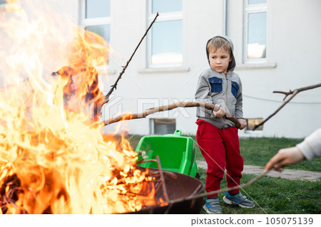 Cute little adorable baby boy put big wooden stick in fire pit with huge fire at house garden yard. Child play dangerous game with bonfire. Fire accident risk and health damgae concept 105075139