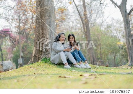 Grown daughter with aging mother expressing love and looking at smartphone mobile under tree in public park 105076867