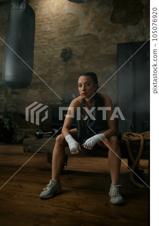 Female boxer in boxing bandage carrying gloves on shoulders sitting on bench 105076920