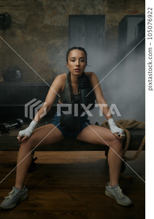 Female boxer in boxing bandage carrying gloves on shoulders sitting on bench 105076922