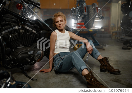 Pretty trendy girl mechanic sitting on garage floor nearby motorcycle 105076954