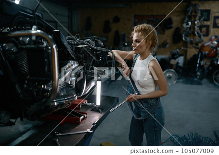 Talented girl wearing blue jumpsuit using work tool to repair motorcycle 105077009