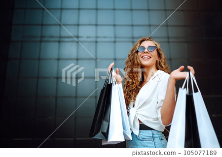 Happy stylish woman with shopping bags enjoys sunny weather walking down street. Summer shopping. Happy stylish woman with shopping bags enjoys sunny weather walking down street. Summer shopping. 105077189