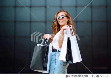 Happy stylish woman with shopping bags enjoys sunny weather walking down street. Summer shopping. 105077195
