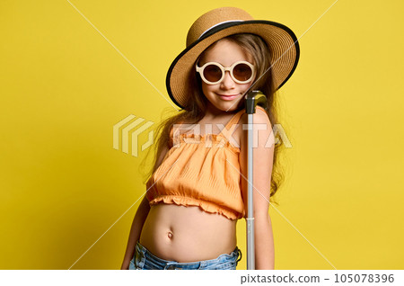 Cute kid girl tourist in straw hat, sunglasses and summer clothes, smiles at camera, travelling abroad, yellow backdrop Cute kid girl tourist in straw hat, sunglasses and summer clothes, smiles at camera, travelling abroad, yellow backdrop 105078396